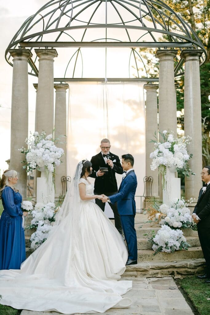 Bride and groom exchanging vows under a domed ceremony arbor styled with white floral arrangements