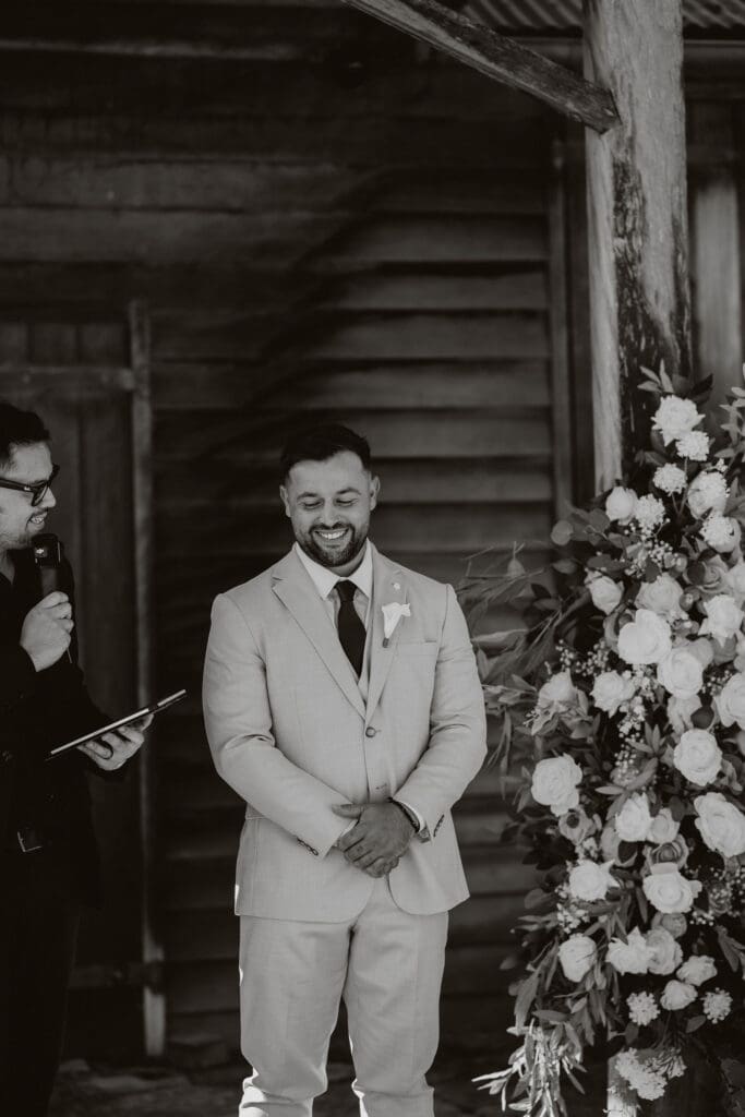 Groom smiling during his wedding ceremony beside a white floral arbor at a barn wedding venue